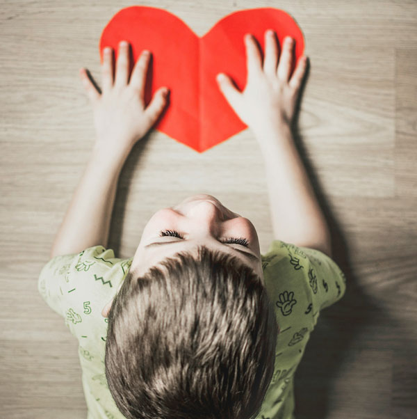 a top-down view of a boy at a table with two hands on a red paper heart