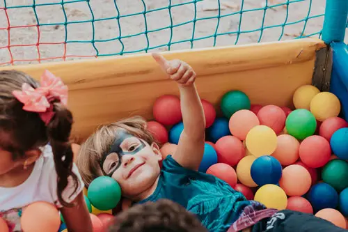 A small boy with a face-painted eye mask lying in a ball pit holding a thumbs up