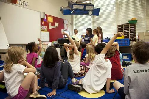 A classroom full of children sitting on a rug, some with raised hands